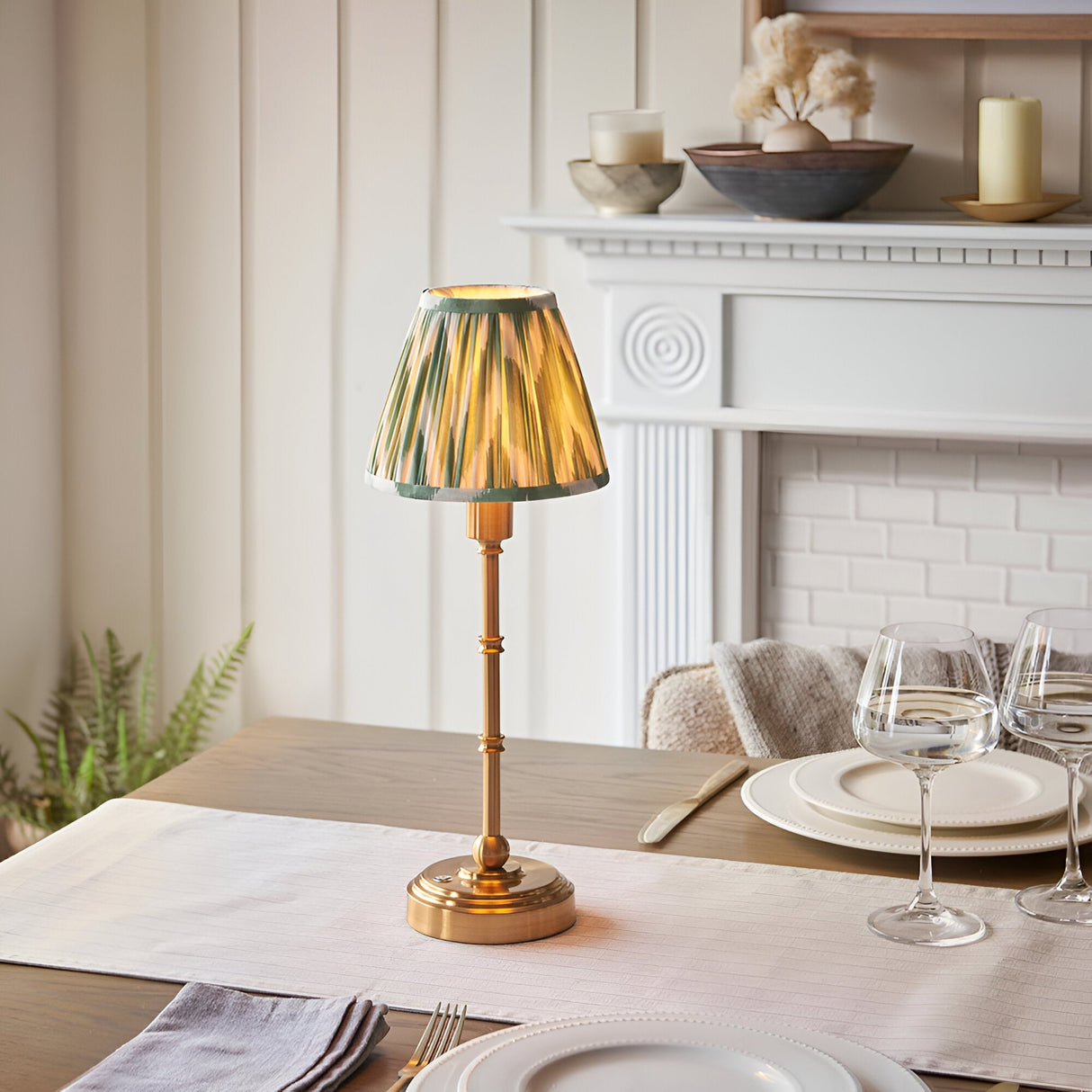 Ashstead LED Zigzag Table Lamp with brass base and green-trimmed patterned fabric shade, shown lit on a dining table near a white fireplace mantel.