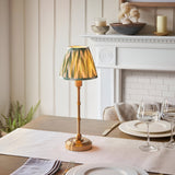 Ashstead LED Zigzag Table Lamp with brass base and green-trimmed patterned fabric shade, shown lit on a dining table near a white fireplace mantel.