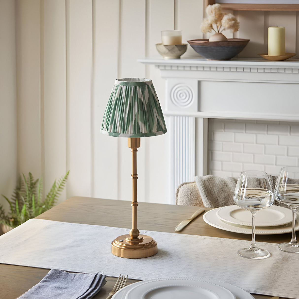 Ashstead LED Zigzag Table Lamp with brass stick base and green-white pleated shade, styled on a dining table beside a white fireplace.