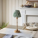 Ashstead LED Zigzag Table Lamp with brass stick base and green-white pleated shade, styled on a dining table beside a white fireplace.
