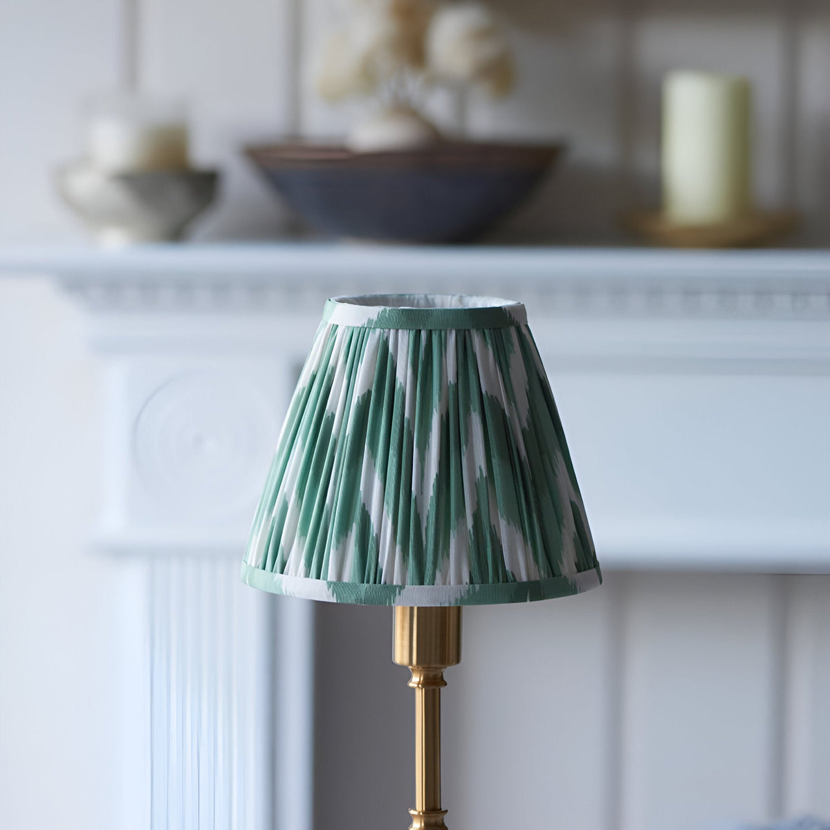 Ashstead LED Zigzag Table Lamp with pleated green and white shade on a brass base, set on a mantel in front of a white panelled fireplace.