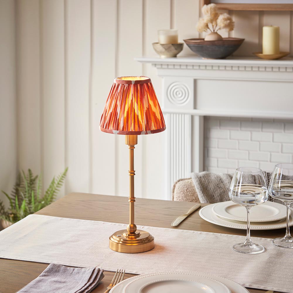 Endon Ashstead cordless LED table lamp with brushed brass stem and pleated apricot zigzag shade, set on a dining table beside wine glasses and plates.