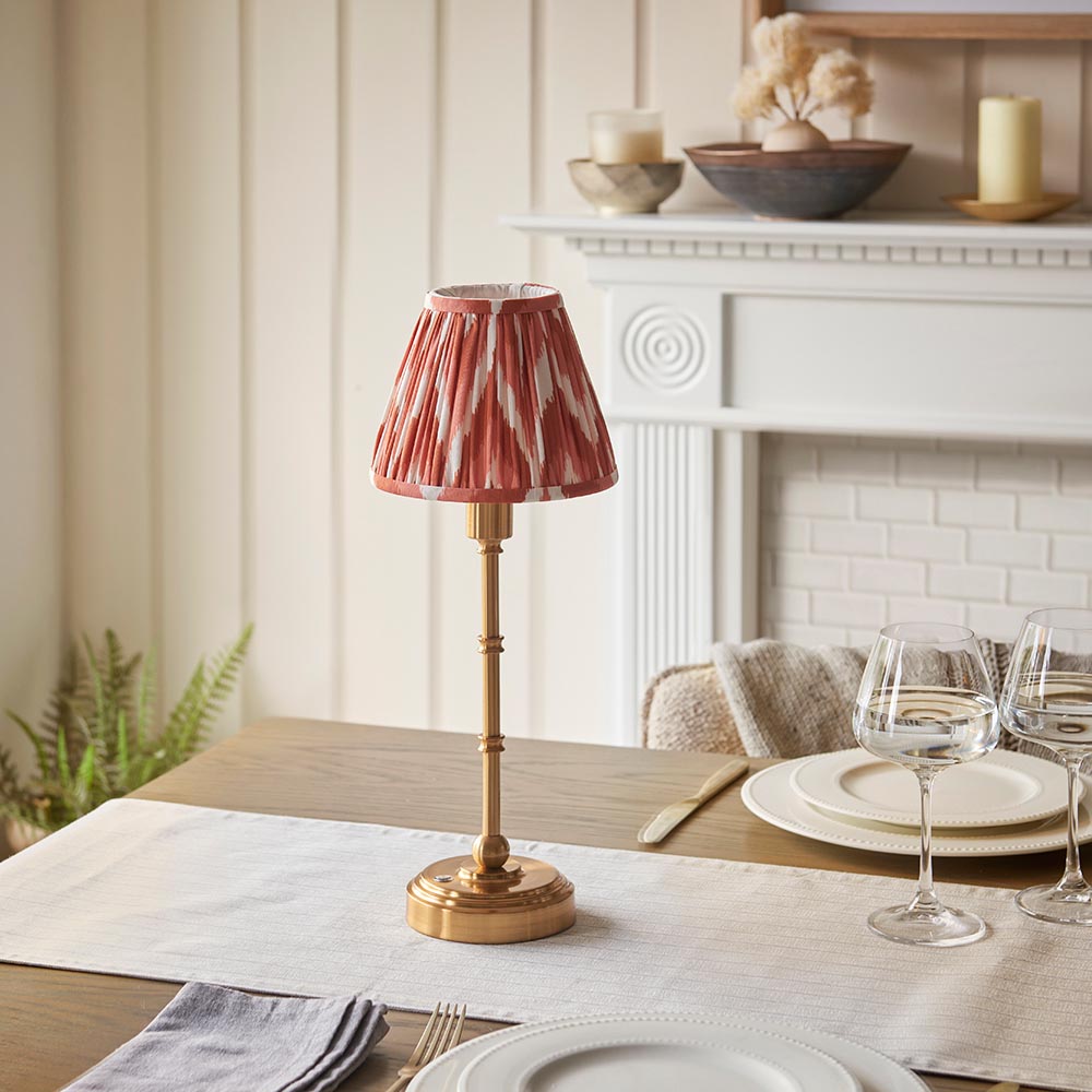Endon Ashstead cordless LED table lamp with brushed brass stem and apricot orange zigzag pleated shade, set on a dining table beside wine glasses and plates.
