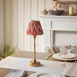 Endon Ashstead cordless LED table lamp with brushed brass stem and apricot orange zigzag pleated shade, set on a dining table beside wine glasses and plates.