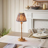Ashstead LED table lamp with brass stem and base, patterned Ikat clip-on shade in brown and grey, shown lit on a dining table near a white fireplace.