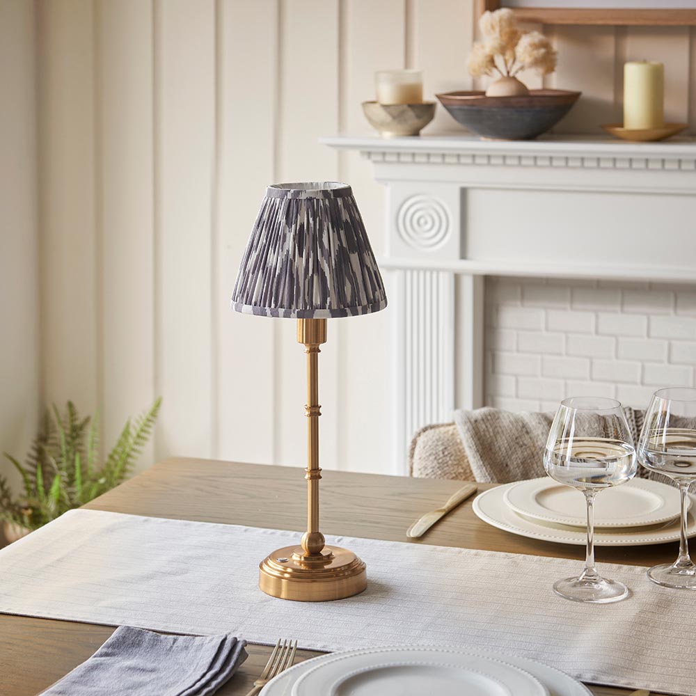 Ashstead LED table lamp with brass stem and base, pleated grey and white Ikat print shade, styled on a dining table near a white fireplace.