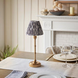 Ashstead LED table lamp with brass stem and base, pleated grey and white Ikat print shade, styled on a dining table near a white fireplace.