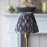 Ashstead LED table lamp with pleated grey and white Ikat fabric shade on a brass base, shown on a mantelpiece in a living room setting.