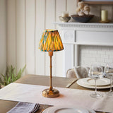 Ashstead LED table lamp with brass base and stem, topped with a multicolour Ikat fabric shade, shown lit on a dining table near a white mantelpiece.
