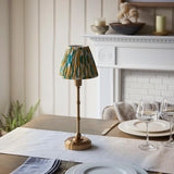 Ashstead LED table lamp with brass stem and base, topped by a yellow, teal and white Ikat fabric shade, shown on a dining table near a fireplace.