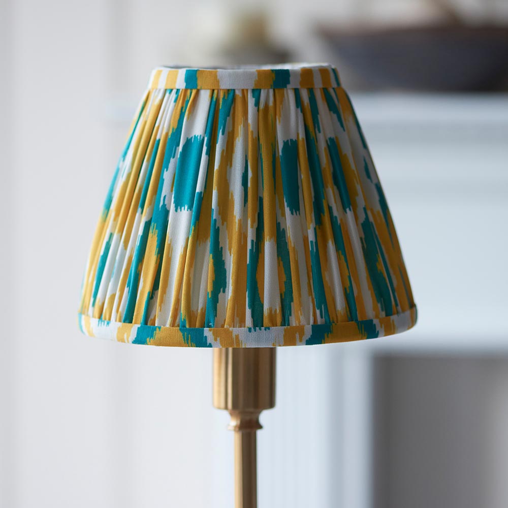 Ashstead LED table lamp with pleated Ikat-print fabric shade in teal, yellow and white on a brass stem, shown close-up against a softly blurred interior.