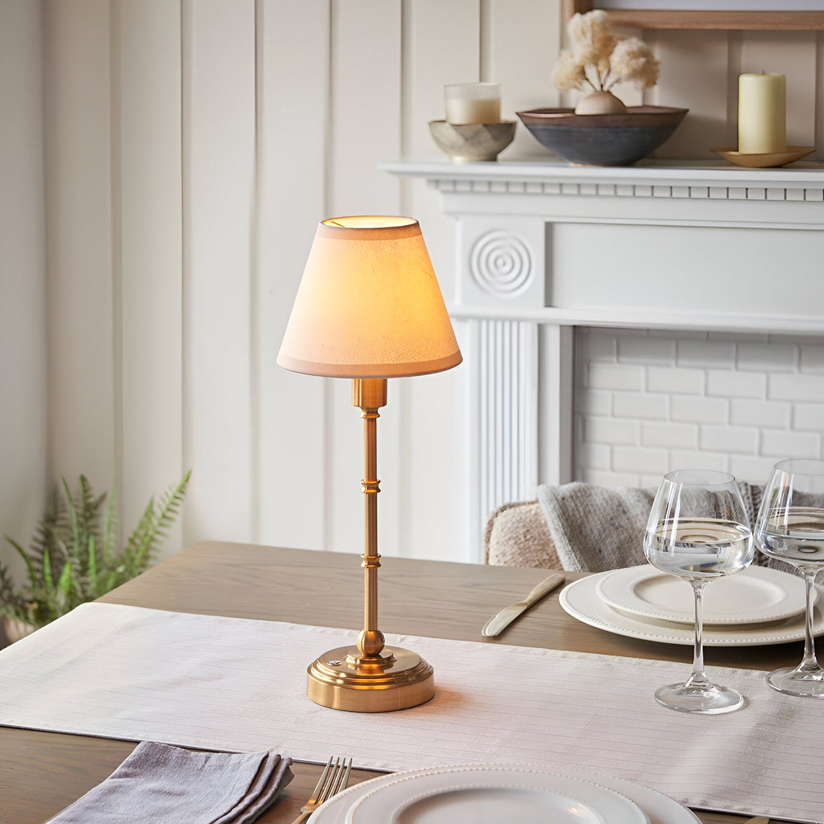 Ashstead LED Table Lamp in brushed aged brass with a vintage white fabric shade, shown lit on a dining table near a white fireplace mantel.