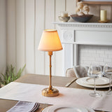 Ashstead LED Table Lamp in brushed aged brass with a vintage white fabric shade, shown lit on a dining table near a white fireplace mantel.