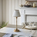 Ashstead LED Table Lamp in brushed aged brass with vintage white fabric shade, styled on a dining table beside a white mantelpiece.