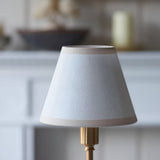 Ashstead LED Table Lamp in vintage white cotton mix tapered shade with brushed brass base, shown close-up against a softly blurred interior backdrop.