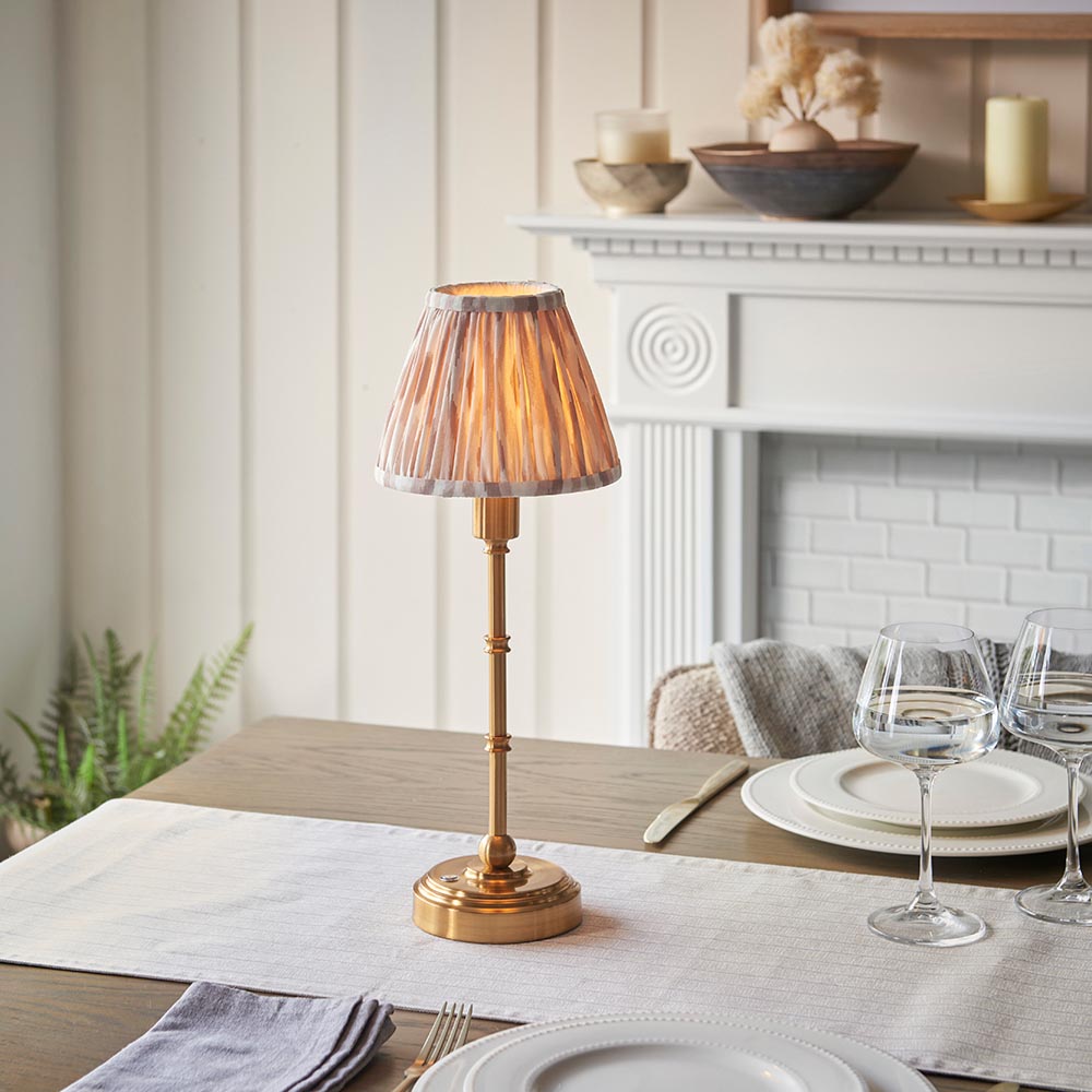Endon Ashstead LED Ikat table lamp in brushed brass with a pleated natural and white shade, set on a dining table beside wine glasses and plates.