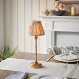 Endon Ashstead LED Ikat table lamp in brushed brass with a pleated natural and white shade, set on a dining table beside wine glasses and plates.