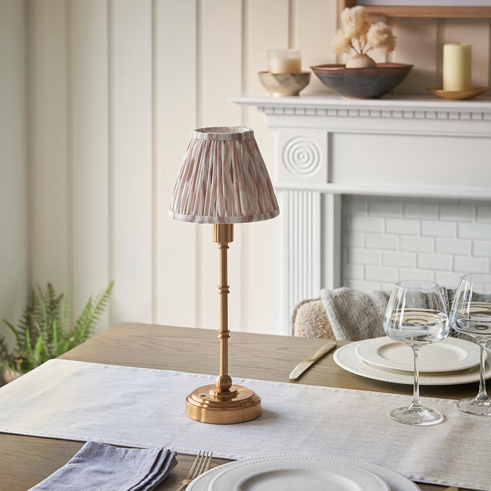 Endon Ashstead LED table lamp with brushed brass stem and pleated Ikat shade in white and natural, on a dining table set with wine glasses and plates.