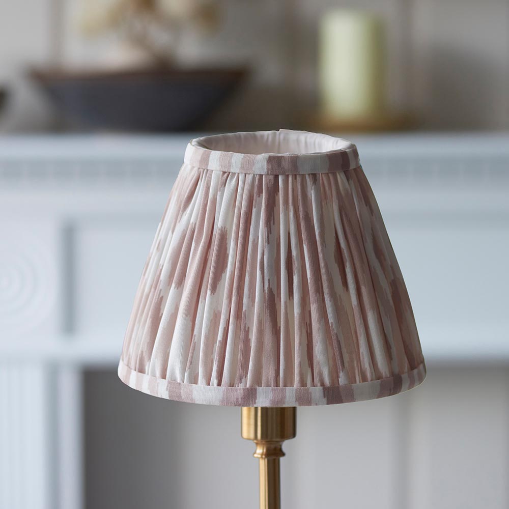 Endon Ashstead LED table lamp with brushed brass base and handmade pleated Ikat clip-on shade in pink and natural tones, shown by a pale mantelpiece.