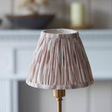 Endon Ashstead LED table lamp with brushed brass base and handmade pleated Ikat clip-on shade in pink and natural tones, shown by a pale mantelpiece.