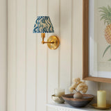 Ashstead LED Ripple wall light with brushed brass arm and blue-and-white patterned fabric shade, mounted beside framed pineapple artwork.