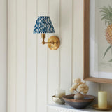 Ashstead LED Ripple wall light with brass arm and blue wave-patterned fabric shade, mounted on a panelled wall above a decorated shelf.