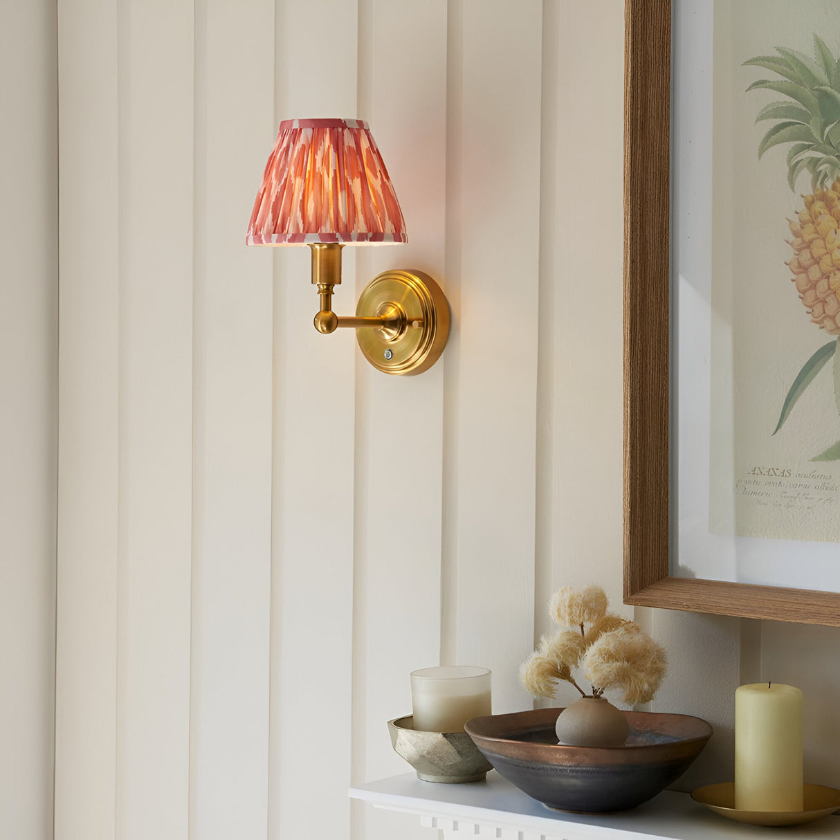 Ashstead LED Ikat wall light with brushed brass arm and pink-and-white patterned fabric shade, shown illuminated on a panelled wall above a decorative shelf.