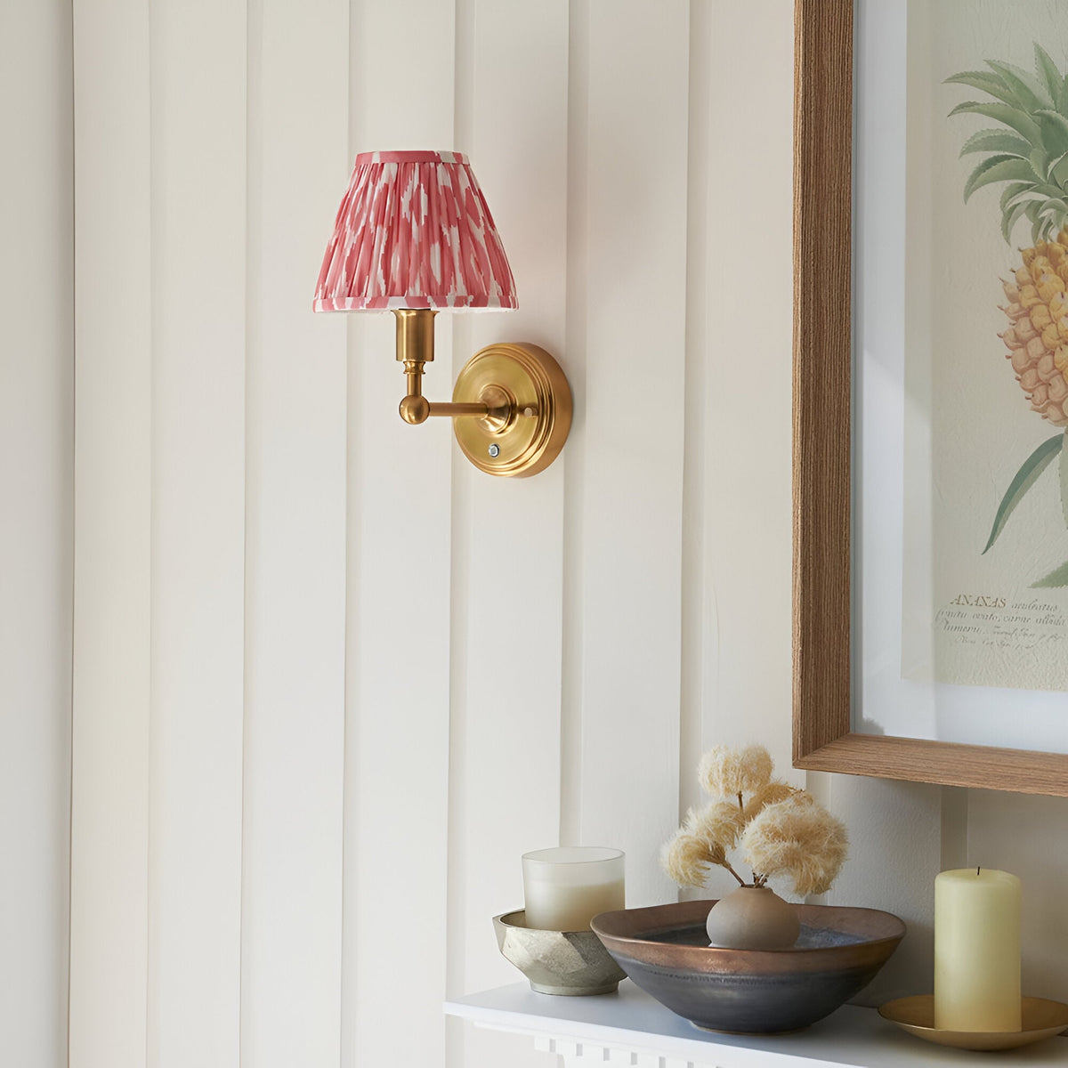 Ashstead LED Ikat wall light with brushed brass arm and pink-and-white pleated shade, mounted beside a framed print and mantel decor.