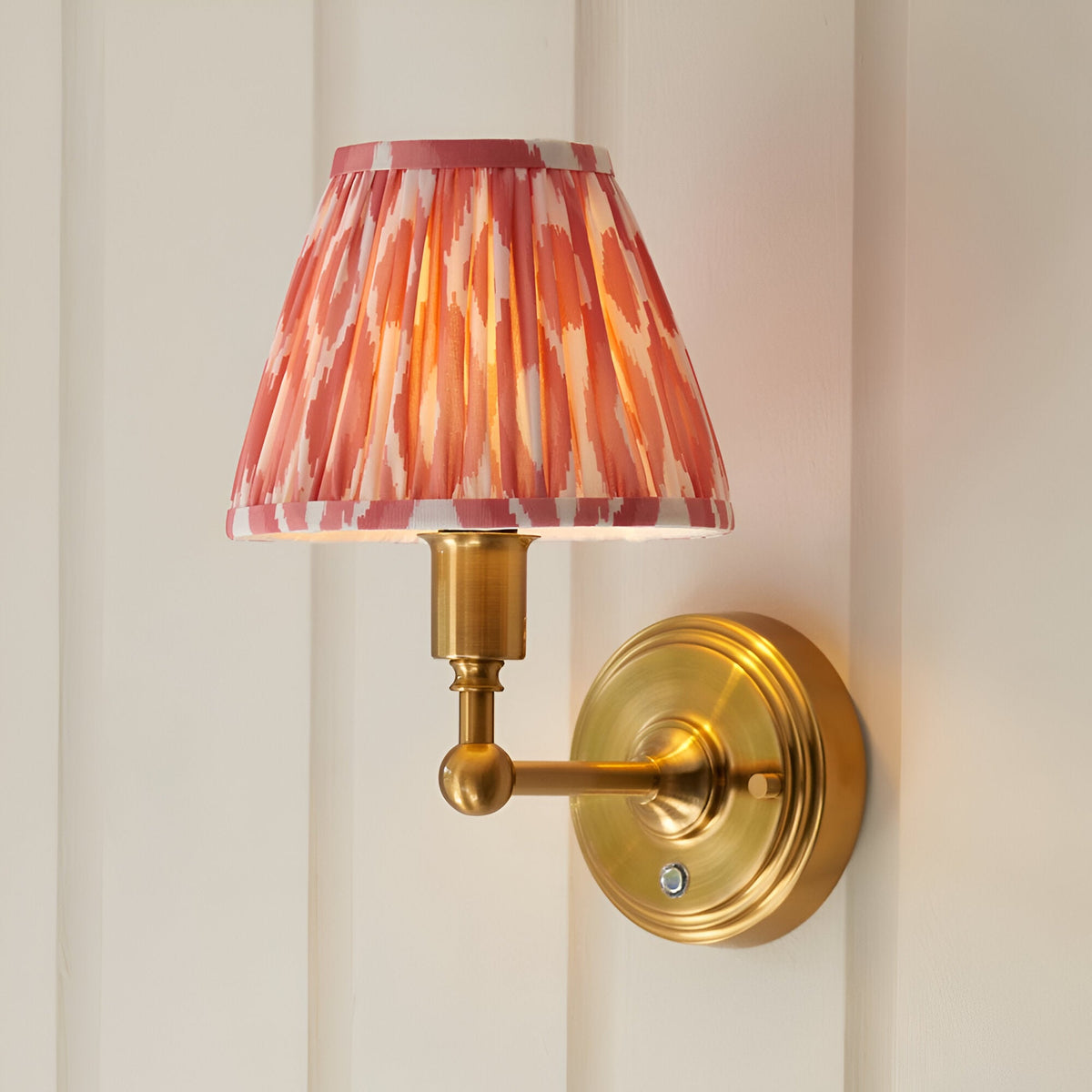 Ashstead LED Ikat wall light with pleated pink and white patterned shade on a brushed brass arm and round wall mount, shown against a light panelled wall.