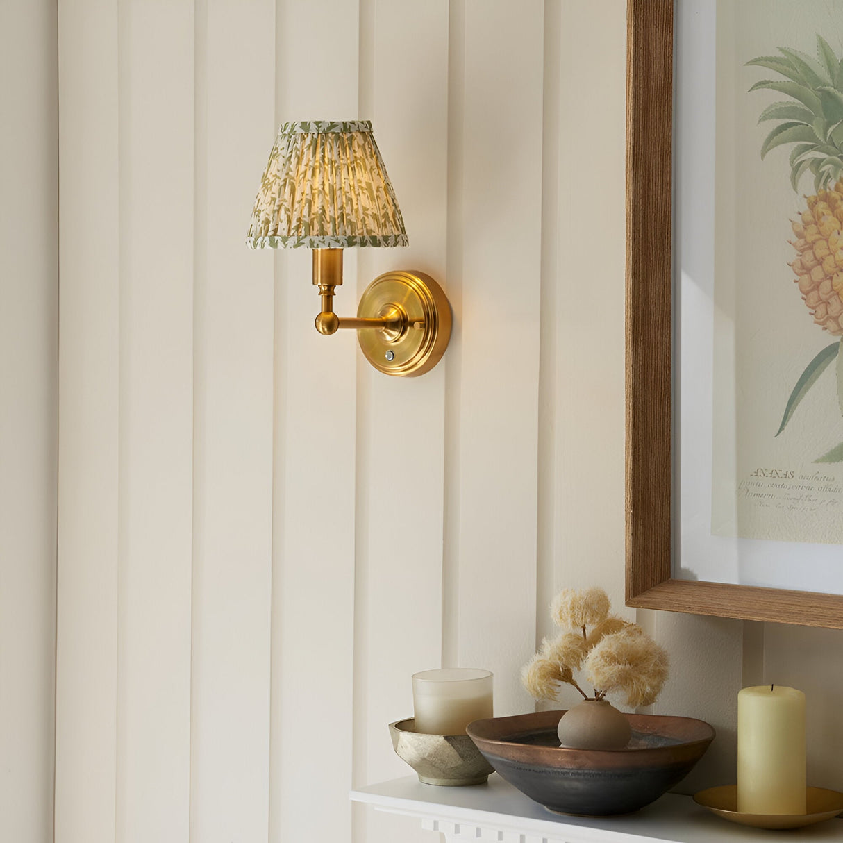 Endon Ashstead LED wall light with brushed brass arm and pleated green leaf-print shade, mounted on cream panelled wall beside a framed pineapple print.