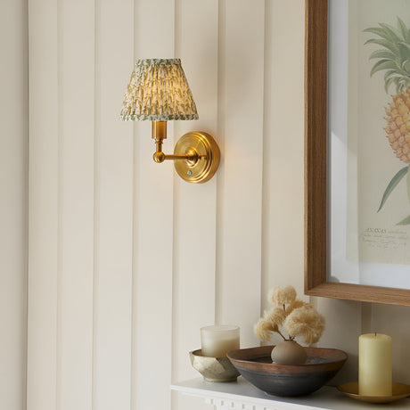 Endon Ashstead LED wall light with brushed brass arm and pleated green leaf-print shade, mounted on cream panelled wall beside a framed pineapple print.