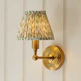Endon Ashstead LED wall light with brushed brass arm and pleated faux linen shade in herb green leaf print, mounted on a cream panelled wall.