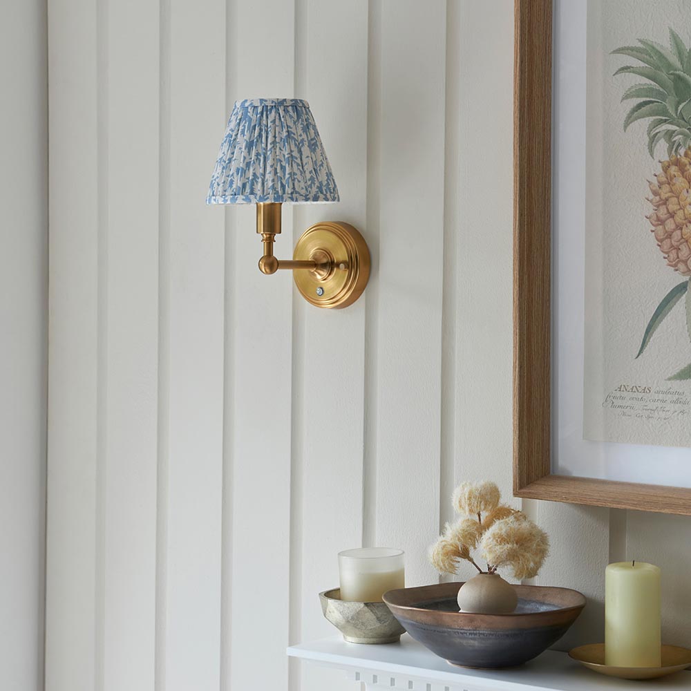 Endon Ashstead LED Leaf wall light with pleated blue and white faux linen shade on a brushed aged brass arm, mounted on panelled wall above decor.