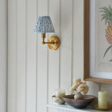 Endon Ashstead LED Leaf wall light with pleated blue and white faux linen shade on a brushed aged brass arm, mounted on panelled wall above decor.