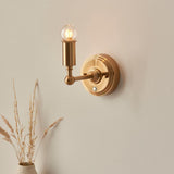 Endon Ashstead LED wall light with a lit globe bulb on a brushed brass arm and round backplate, mounted on a neutral wall above dried grasses.
