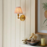 Ashstead LED Leaf Wall Light in brass with a pleated fabric shade, mounted on a panelled wall above a shelf with decor.