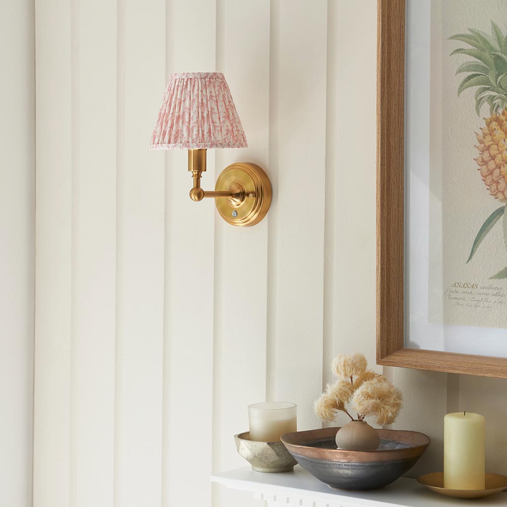 Ashstead LED Leaf wall light with pleated pink patterned fabric shade on a brushed brass arm, mounted on a panelled cream wall above a decorative shelf.