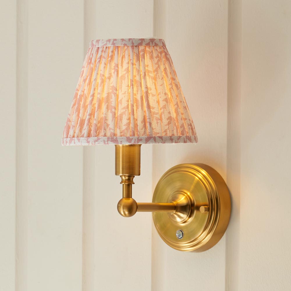 Ashstead LED Leaf wall light with pleated pink and cream patterned shade on a brushed brass arm and round wall plate, mounted on a white panelled wall.