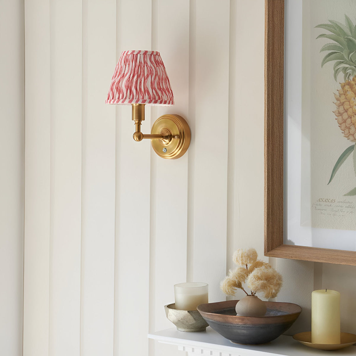 Endon Ashstead LED Ripple wall light with brushed brass arm and pink and white pleated fabric shade, mounted on a panelled wall beside a framed pineapple print.