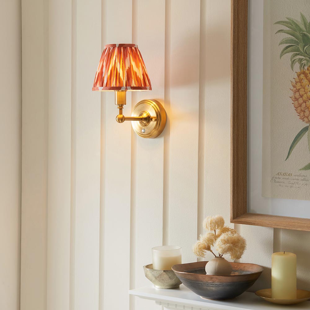 Ashstead LED Zigzag Wall Light with brass arm and orange patterned fabric shade, mounted on a panelled wall above a hallway console with decor.