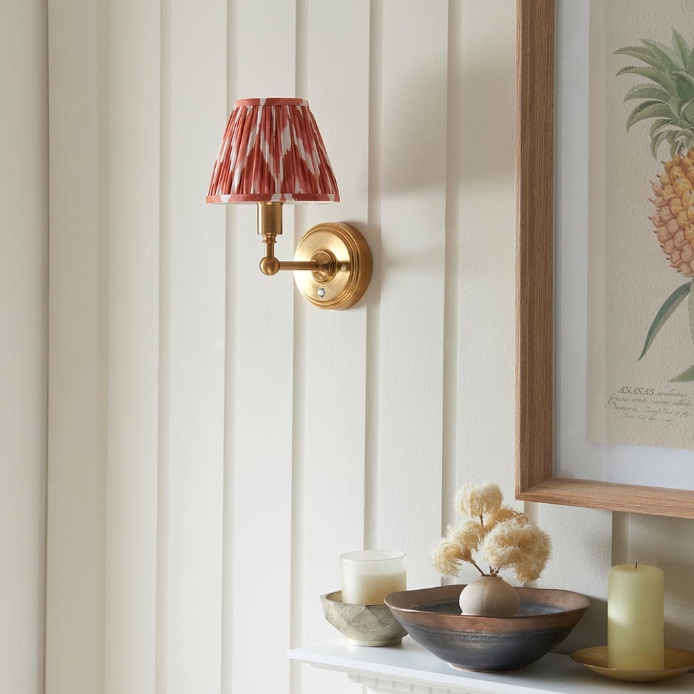 Ashstead LED Zigzag wall light with brass arm and red-and-white patterned fabric shade, mounted on a panelled wall above a decorative console shelf.