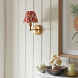 Ashstead LED Zigzag wall light with brass arm and red-and-white patterned fabric shade, mounted on a panelled wall above a decorative console shelf.