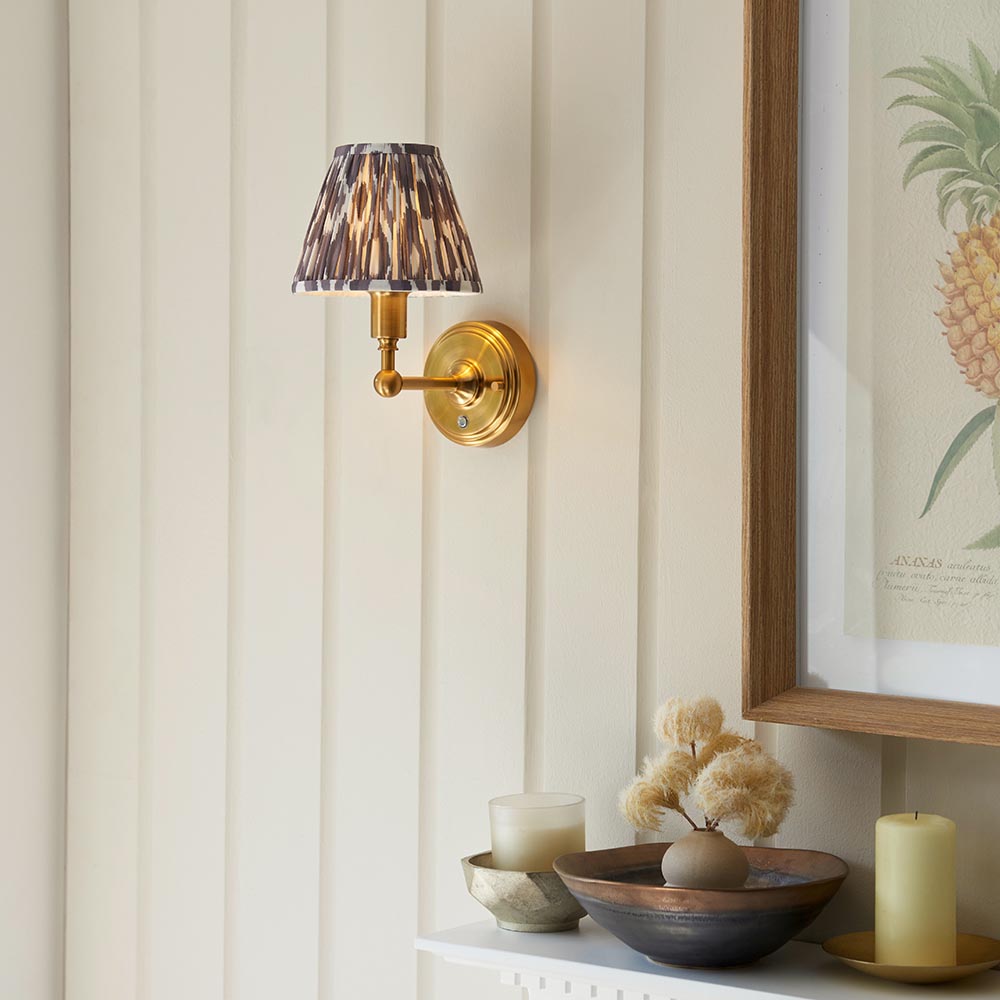 Ashstead LED Ikat wall light with brushed brass arm and patterned fabric shade, mounted on a panelled wall above a shelf styled with candles and a framed print.