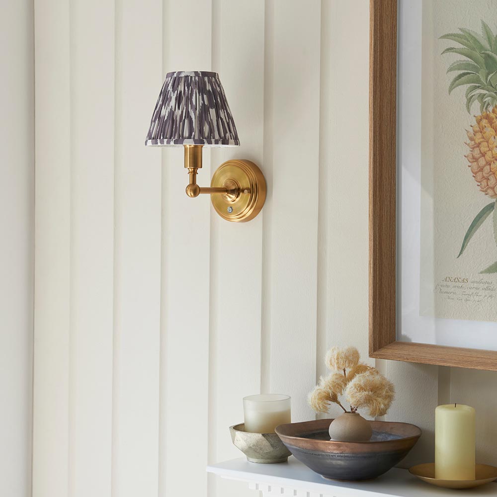 Ashstead LED Ikat wall light with brushed brass arm and patterned grey and white fabric shade, mounted on a panelled wall near a shelf display.