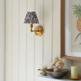 Ashstead LED Ikat wall light with brushed brass arm and patterned grey and white fabric shade, mounted on a panelled wall near a shelf display.