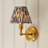 Ashstead LED Ikat wall light with pleated brown, white and grey shade on a brushed brass arm, mounted on a light panelled wall.