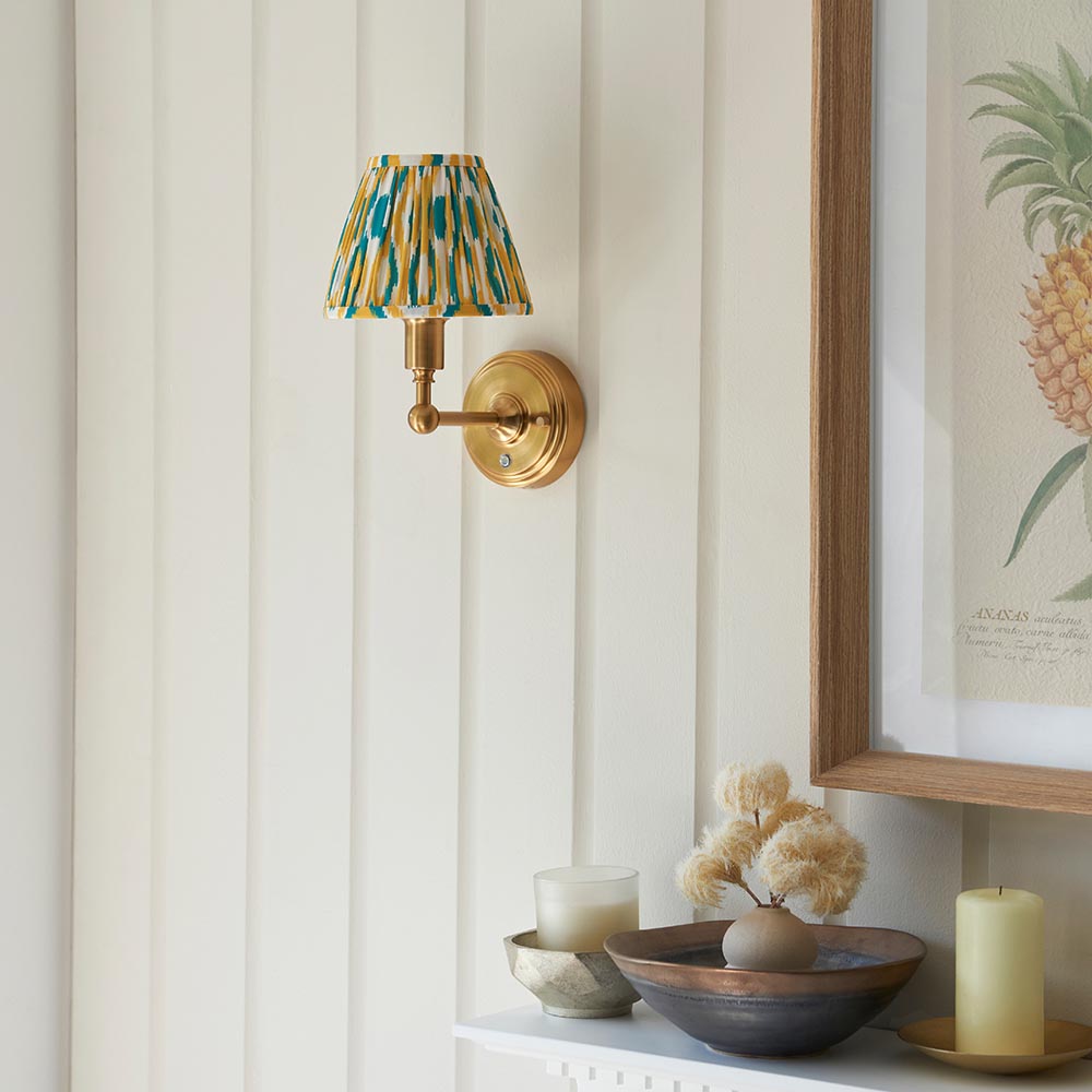 Endon Ashstead LED Ikat wall light with brushed brass arm and pleated white, yellow and teal ikat shade, mounted beside a framed pineapple print.