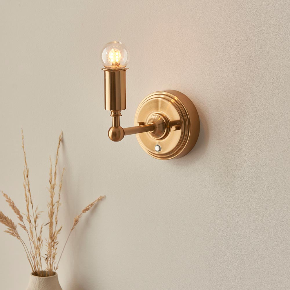 Endon Ashstead LED wall light with brushed brass arm and lit globe bulb, mounted on a neutral wall beside dried grasses in a ceramic vase.