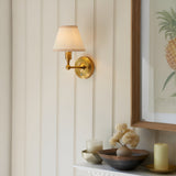 Endon Ashstead LED rechargeable wall light in brushed aged brass with tapered ivory fabric shade, mounted on panelled wall beside framed pineapple print.