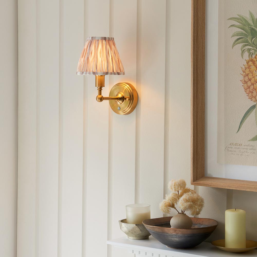 Endon Ashstead LED wall light with brushed brass arm and pleated white fabric shade, lit on a panelled wall beside a framed pineapple print.